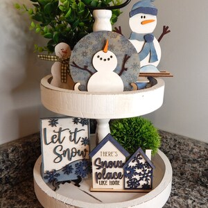 May include: A white tiered tray with a winter theme. The top tier features a blue and white snowman ornament, a blue snowman figurine, and a green bush. The bottom tier has a sign that says "Let it Snow" and a sign that says "There's Snow Place Like Home".
