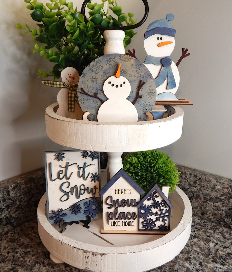 May include: A white tiered tray with a winter theme. The top tier features a blue and white snowman ornament, a blue snowman figurine, and a green bush. The bottom tier has a sign that says "Let it Snow" and a sign that says "There's Snow Place Like Home".