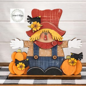 May include: A wooden scarecrow decoration featuring a red hat, yellow hair, and blue overalls. The figure is flanked by orange pumpkins, black birds, and yellow flowers with brown button centers. The design includes white polka dots and cross-hatch details.