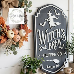 May include: A dark gray, shield-shaped wooden sign with white 3D lettering that reads Witch's Brew Coffee Co. Salem MA. The sign features a white silhouette of a witch on a broomstick, three bats, and a broom icon. It is displayed next to a fall wreath.