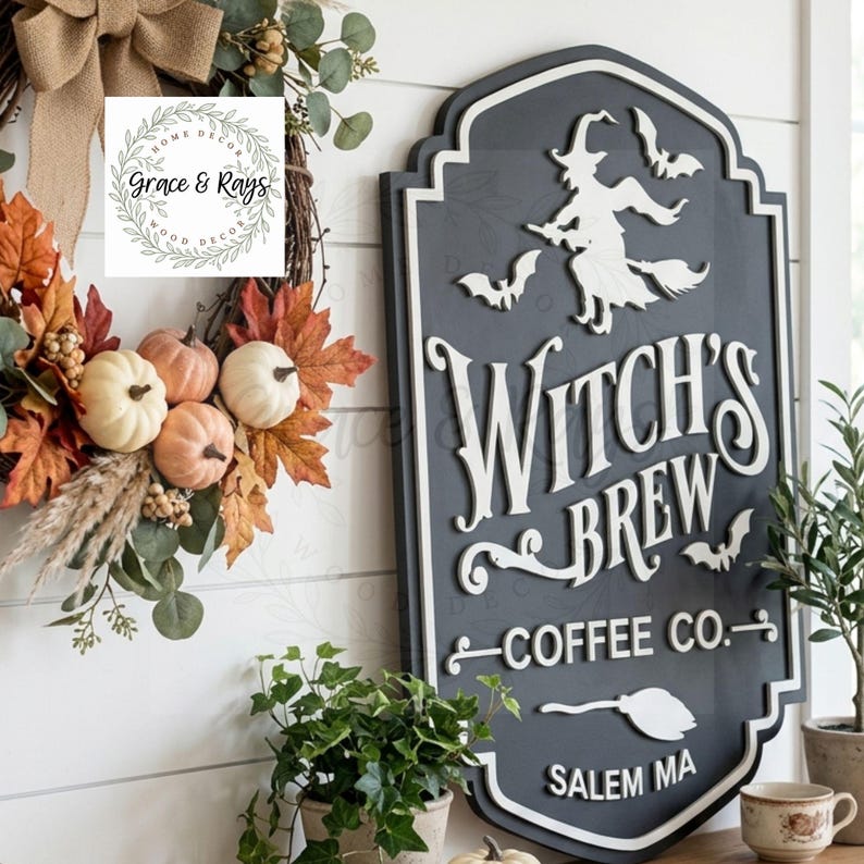 May include: A dark gray, shield-shaped wooden sign with white 3D lettering that reads Witch's Brew Coffee Co. Salem MA. The sign features a white silhouette of a witch on a broomstick, three bats, and a broom icon. It is displayed next to a fall wreath.