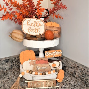 May include: A two-tiered white tiered tray decorated with autumn-themed wooden signs and decorations. The top tier features a wooden pumpkin sign that says "hello fall" and a small wooden crate filled with miniature pumpkins. The bottom tier features a wooden sign that says "pumpkins", a wooden sign that says "grateful", and a wooden sign that says "pumpkin patch". The tray is decorated with a garland of orange and white beads and a brown tassel.