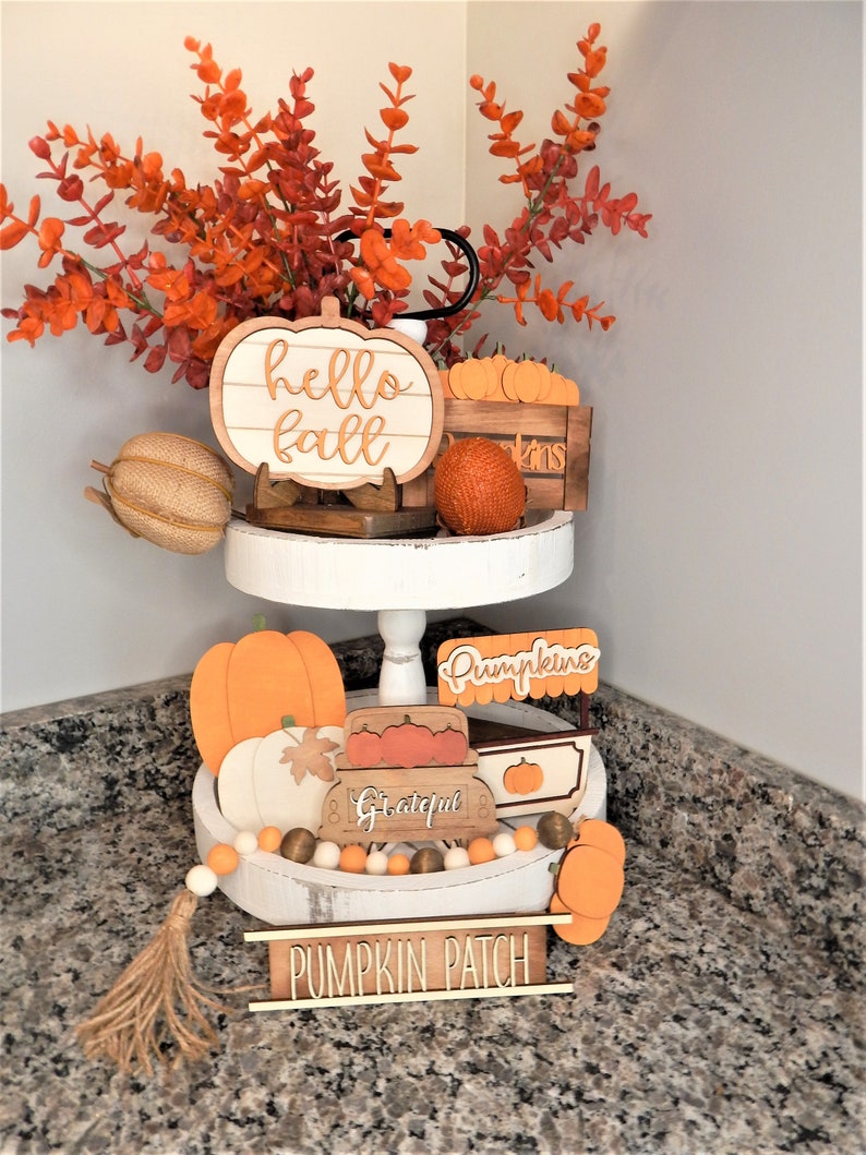 May include: A two-tiered white tiered tray decorated with autumn-themed wooden signs and decorations. The top tier features a wooden pumpkin sign that says "hello fall" and a small wooden crate filled with miniature pumpkins. The bottom tier features a wooden sign that says "pumpkins", a wooden sign that says "grateful", and a wooden sign that says "pumpkin patch". The tray is decorated with a garland of orange and white beads and a brown tassel.