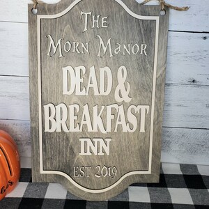 May include: A wooden sign with a distressed gray finish and white lettering. The sign reads "The Morn Manor Dead & Breakfast Inn Est 2019".
