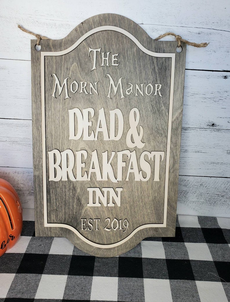 May include: A wooden sign with a distressed gray finish and white lettering. The sign reads "The Morn Manor Dead & Breakfast Inn Est 2019".