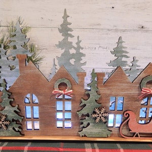 May include: A wooden Christmas village scene with three houses, a sleigh, and trees. The houses have windows and wreaths with red and white ribbon. The trees are green and have snowflakes. The scene is lit up with white lights.