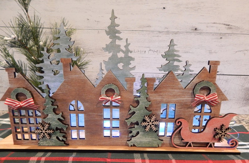 May include: A wooden Christmas village scene with three houses, a sleigh, and trees. The houses have windows and wreaths with red and white ribbon. The trees are green and have snowflakes. The scene is lit up with white lights.