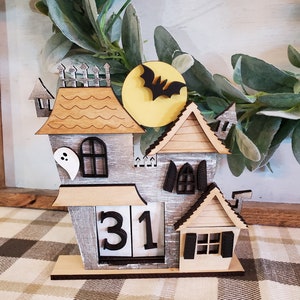 Puede incluir: Un calendario de Halloween de madera con forma de casa embrujada. La casa es gris con detalles de madera, un fantasma y una silueta de murci&eacute;lago frente a una luna amarilla. El calendario muestra el n&uacute;mero 31.