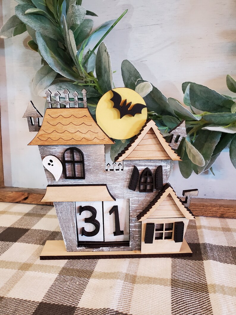 Puede incluir: Un calendario de Halloween de madera con forma de casa embrujada. La casa es gris con detalles de madera, un fantasma y una silueta de murci&eacute;lago frente a una luna amarilla. El calendario muestra el n&uacute;mero 31.