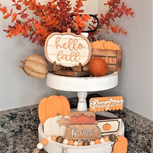 May include: A white tiered tray decorated with autumn-themed wooden signs, pumpkins, and a garland of white and orange beads. The signs read "hello fall", "pumpkins", "grateful", and "pumpkin patch".
