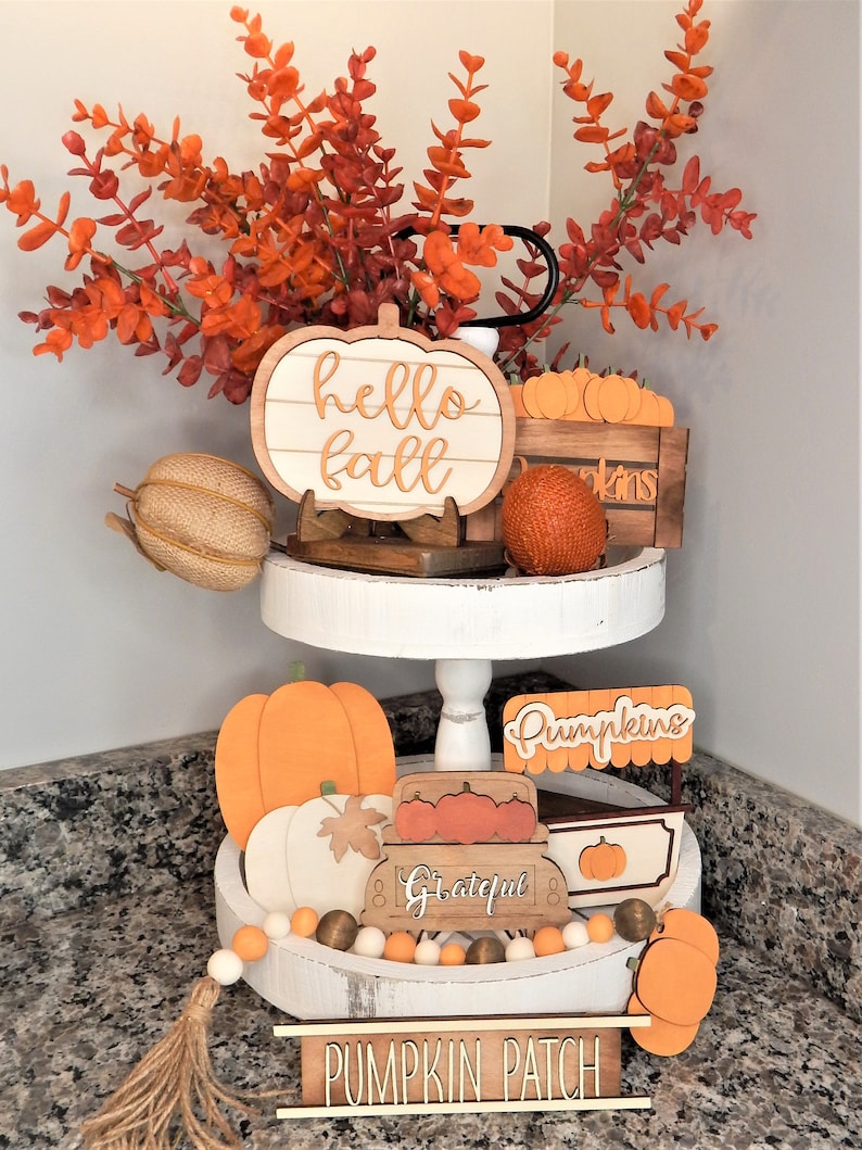May include: A white tiered tray decorated with autumn-themed wooden signs, pumpkins, and a garland of white and orange beads. The signs read "hello fall", "pumpkins", "grateful", and "pumpkin patch".