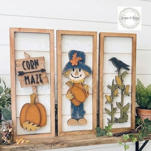 May include: Three rectangular wooden wall signs featuring fall themes. The left sign displays a wooden pumpkin and a sign reading CORN MAZE. The center sign features a scarecrow with a blue hat and pumpkin. The right sign shows a black crow on corn.