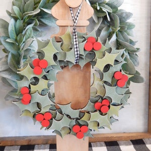 May include: A wooden Christmas wreath with green holly leaves and red berries. The wreath is displayed on a wooden stand with a black and white checkered ribbon. The background features a green leafy wreath.