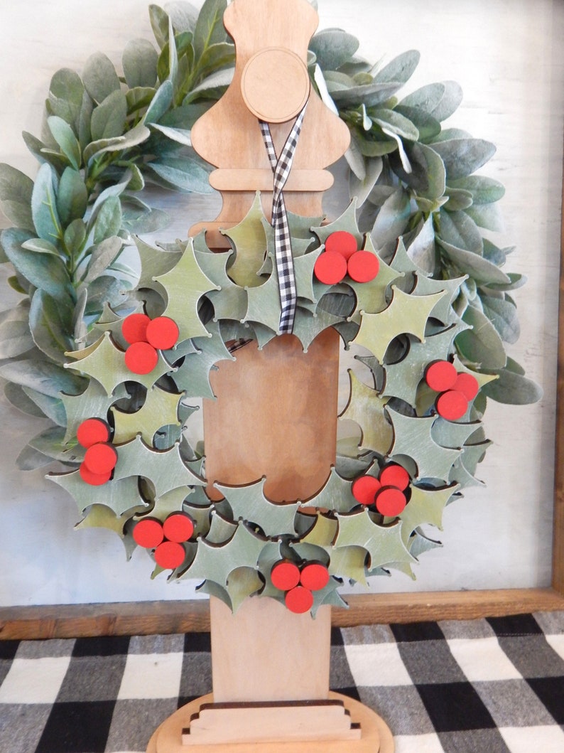 May include: A wooden Christmas wreath with green holly leaves and red berries. The wreath is displayed on a wooden stand with a black and white checkered ribbon. The background features a green leafy wreath.