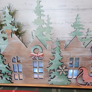 May include: Wooden Christmas village scene with houses, trees, and a Santa sleigh. The houses have windows and wreaths with red and white ribbon. The trees are green and have snowflakes. The sleigh is red and has a white snowflake.