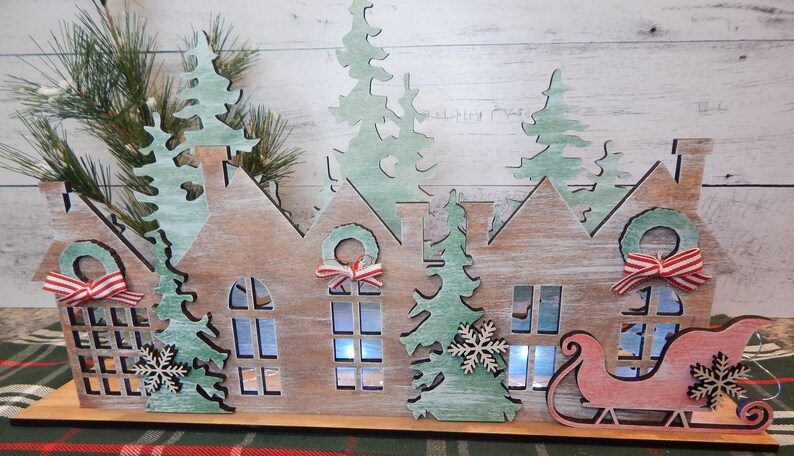 May include: Wooden Christmas village scene with houses, trees, and a Santa sleigh. The houses have windows and wreaths with red and white ribbon. The trees are green and have snowflakes. The sleigh is red and has a white snowflake.