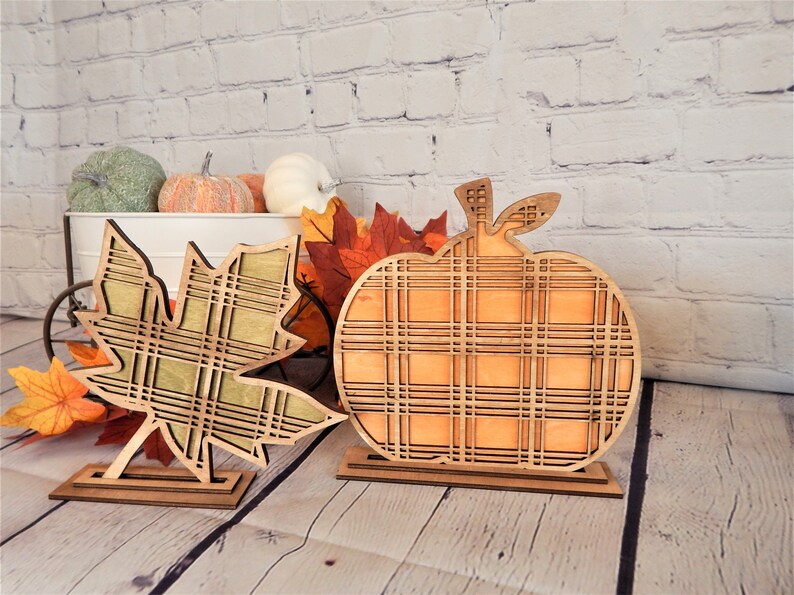 May include: Two wooden fall decorations, a pumpkin and a maple leaf, both with a plaid pattern. The pumpkin is orange and the leaf is green.
