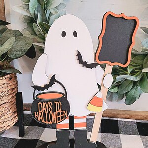 May include: A white ghost holding a cauldron with the text "Days to Halloween" and a candy corn. The ghost is holding a black chalkboard with a white frame. The ghost is standing on a brown wooden base.