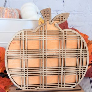 May include: A wooden pumpkin cutout with a plaid pattern. The pumpkin is painted orange and has a brown stem. The pumpkin is sitting on a wooden base.