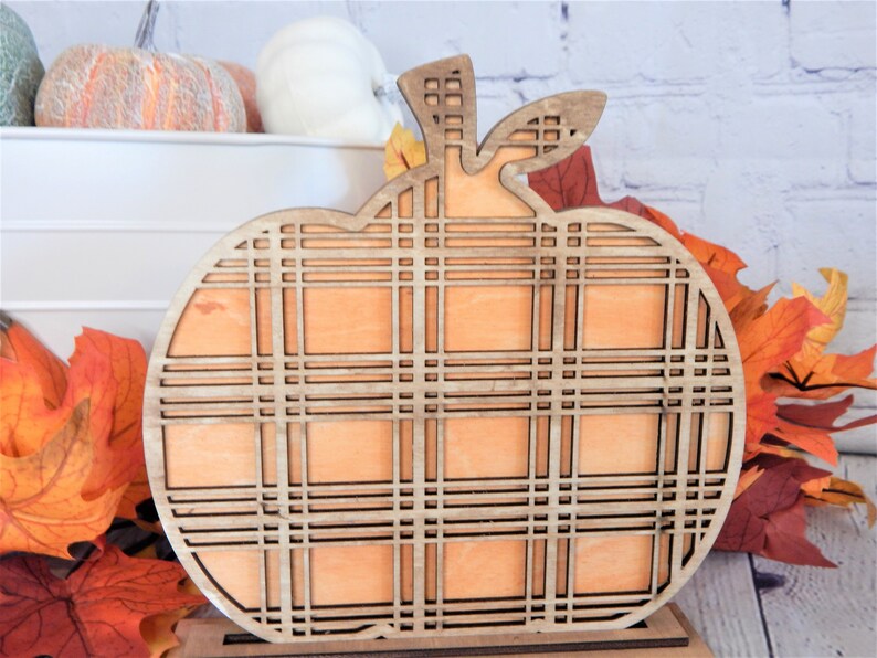 May include: A wooden pumpkin cutout with a plaid pattern. The pumpkin is painted orange and has a brown stem. The pumpkin is sitting on a wooden base.