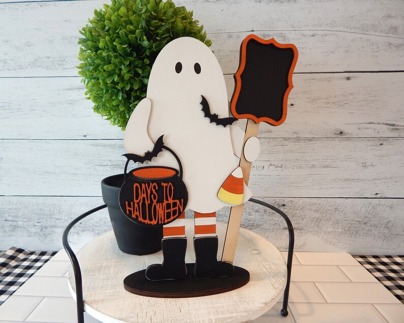 May include: A white ghost figurine holding a black cauldron with the text "Days to Halloween" and a black chalkboard sign. The ghost is wearing orange and white striped stockings and black boots. The figurine is standing on a wooden base.
