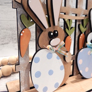 Pu&ograve; includere: Decorazione pasquale in legno con due figure di conigli con corpi a forma di uovo. I conigli sono marroni e bianchi, con uova a pois blu e bianche. Carote e una recinzione fanno parte del design, con nastri pastello.
