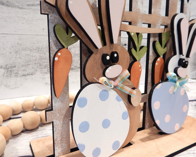 Pu&ograve; includere: Decorazione pasquale in legno con due figure di conigli con corpi a forma di uovo. I conigli sono marroni e bianchi, con uova a pois blu e bianche. Carote e una recinzione fanno parte del design, con nastri pastello.