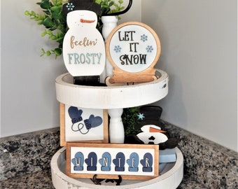 May include: A white tiered tray with winter-themed decorations. The top tier features a snowman with the text "feelin' frosty", a snow globe with the text "Let it snow", and a small green bush. The bottom tier features a sign with the text "WINTER" made of blue mittens, a snowman, and a pair of blue mittens.