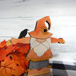 May include: A wooden cutout of a scarecrow with a sunflower on its hat. The scarecrow is wearing a blue shirt and brown pants. The scarecrow is standing on a wooden base. There are fall leaves in the background.