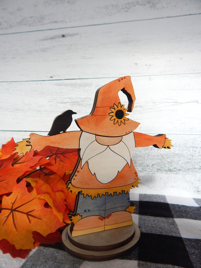 May include: A wooden cutout of a scarecrow with a sunflower on its hat. The scarecrow is wearing a blue shirt and brown pants. The scarecrow is standing on a wooden base. There are fall leaves in the background.