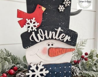 May include: A wooden snowman decoration featuring a black top hat with white speckles, a red cardinal, and white snowflakes. The word Winter is written in white script across the hat brim. The snowman has a carrot nose and pink cheeks.