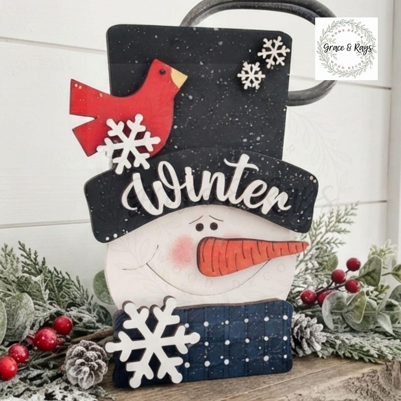 May include: A wooden snowman decoration featuring a black top hat with white speckles, a red cardinal, and white snowflakes. The word Winter is written in white script across the hat brim. The snowman has a carrot nose and pink cheeks.