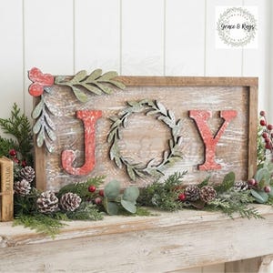 May include: A rectangular wooden sign with a distressed white finish featuring the word JOY in red, where the letter O is replaced by a green leaf wreath. A decorative red and green sprig is attached to the top left corner of the wooden frame.