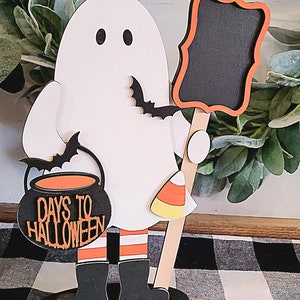 May include: A white ghost holding a cauldron with the text "Days to Halloween" and a candy corn. The ghost is holding a black chalkboard with a red frame. The ghost is standing on a wooden base.