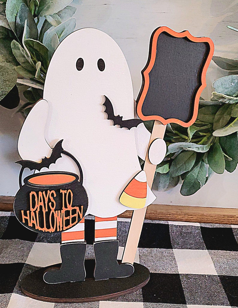 May include: A white ghost holding a cauldron with the text "Days to Halloween" and a candy corn. The ghost is holding a black chalkboard with a red frame. The ghost is standing on a wooden base.