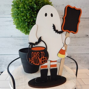 May include: A white ghost holding a black cauldron with the text "Days to Halloween" in orange. The ghost is wearing black boots with orange and white stripes and is holding a black chalkboard with a wooden stick. The ghost is standing on a black base.