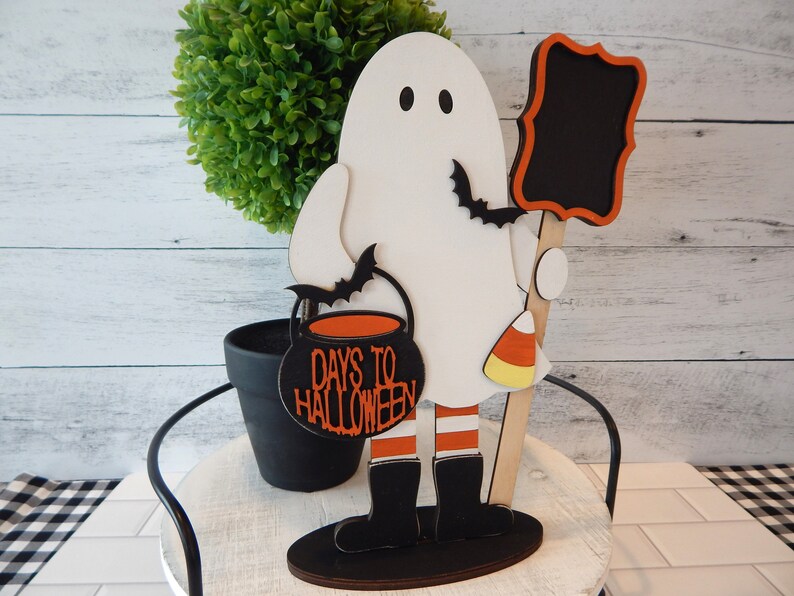 May include: A white ghost holding a black cauldron with the text "Days to Halloween" in orange. The ghost is wearing black boots with orange and white stripes and is holding a black chalkboard with a wooden stick. The ghost is standing on a black base.