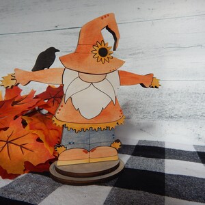May include: A wooden cutout of a scarecrow gnome wearing a hat with a sunflower design. The gnome is holding a small black bird in its hand. The gnome is standing on a wooden base and is surrounded by fall leaves.