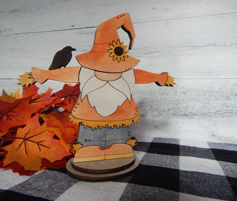 May include: A wooden cutout of a scarecrow gnome wearing a hat with a sunflower design. The gnome is holding a small black bird in its hand. The gnome is standing on a wooden base and is surrounded by fall leaves.