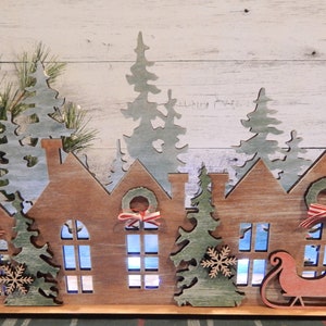 May include: Wooden Christmas village scene with houses, trees, and a Santa sleigh. The houses have windows and wreaths with red and white ribbon. The trees are green and white. The sleigh is red and white.
