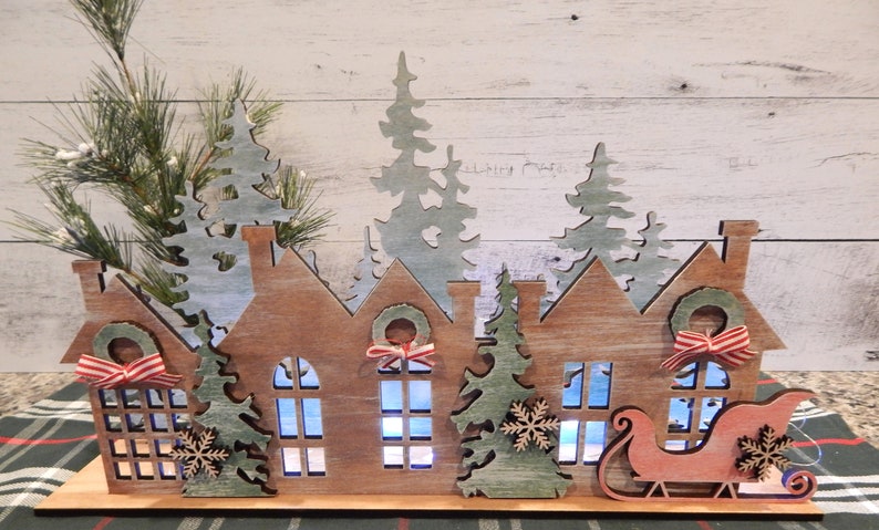 May include: Wooden Christmas village scene with houses, trees, and a Santa sleigh. The houses have windows and wreaths with red and white ribbon. The trees are green and white. The sleigh is red and white.