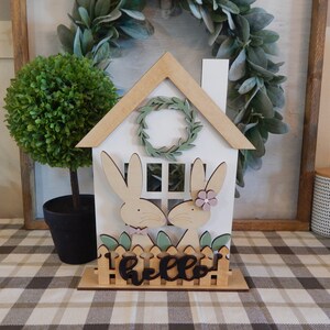 May include: Wooden house shaped decoration with two bunny rabbits peeking out of the windows. The house has a white base with a brown roof and a wreath above the door. The bunnies are painted in pink and brown with a green bush behind them. The words "hello" are written in black on a wooden fence at the bottom of the house.