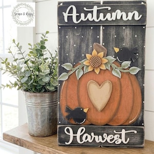 May include: A rectangular wooden sign with a dark distressed finish featuring the word Autumn at the top and Harvest at the bottom in white script. A large orange pumpkin with a central heart shape, a sunflower, and two black birds.
