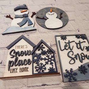 May include: Four wooden Christmas decorations with a winter theme. Two snowmen, one with a blue scarf and one without, and two signs. One sign says "There's snow place like home" and the other says "Let it snow".