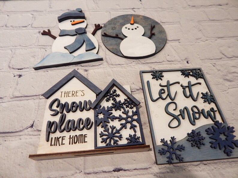 May include: Four wooden Christmas decorations with a winter theme. Two snowmen, one with a blue scarf and one without, and two signs. One sign says "There's snow place like home" and the other says "Let it snow".