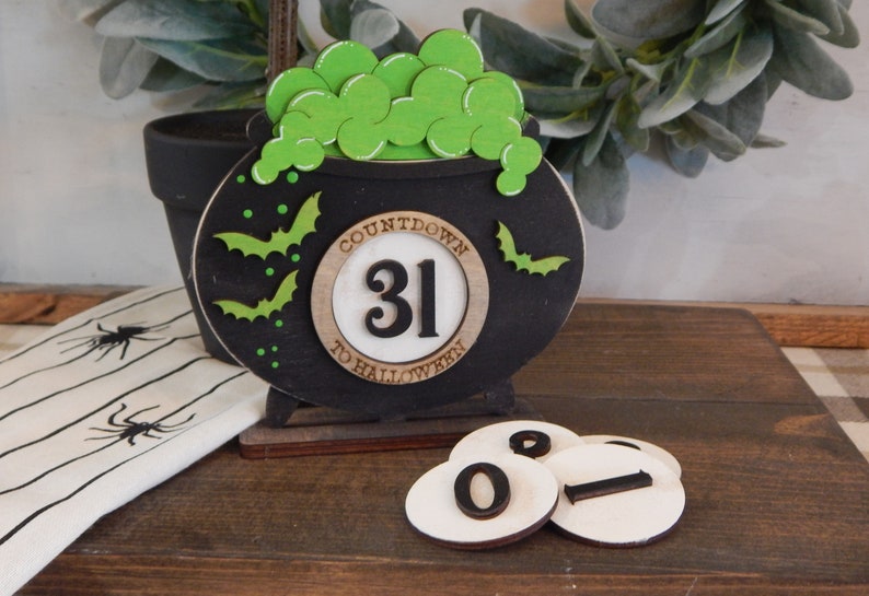 Pu&ograve; includere: Decorazione a conto alla rovescia di Halloween in legno nero a forma di calderone, con bolle e pipistrelli verdi. Il numero 31 &egrave; visualizzato, con tessere numeriche aggiuntive. Il testo "COUNTDOWN TO HALLOWEEN" &egrave; visibile.