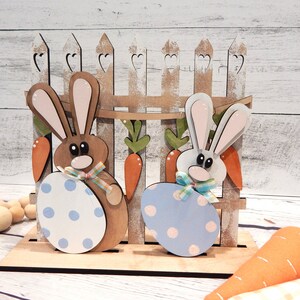 Pu&ograve; includere: Decorazione pasquale in legno con due figure di coniglietti che tengono uova a pois. I coniglietti sono davanti a una staccionata di legno con dettagli di carote e foglie. Una carota di feltro &egrave; in primo piano. I colori sono marrone, bianco, arancione e blu.