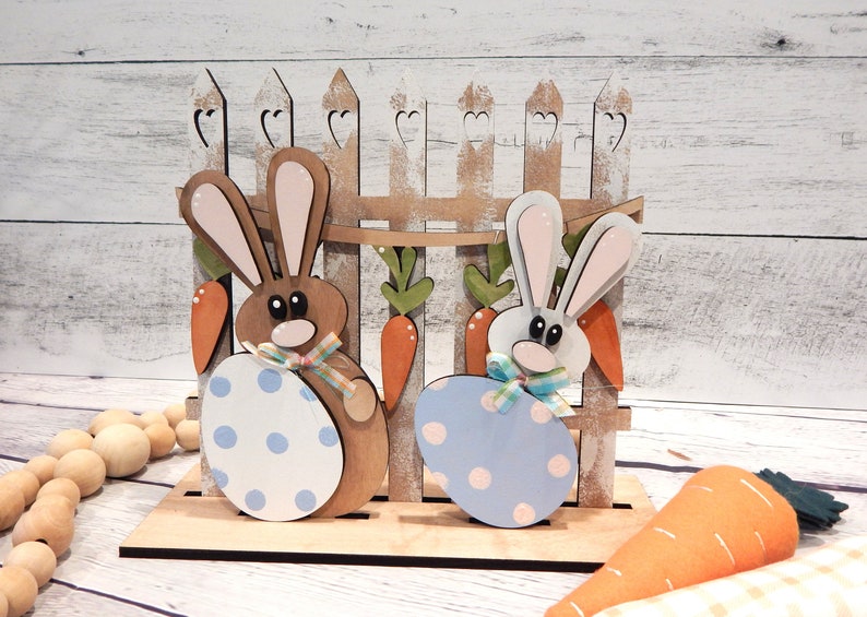 Pu&ograve; includere: Decorazione pasquale in legno con due figure di coniglietti che tengono uova a pois. I coniglietti sono davanti a una staccionata di legno con dettagli di carote e foglie. Una carota di feltro &egrave; in primo piano. I colori sono marrone, bianco, arancione e blu.