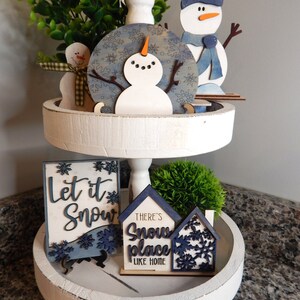 May include: A white tiered tray decorated with winter-themed wooden cutouts. The top tier features a snowman, a blue and white snowflake design, and a small green bush. The bottom tier features a sign that says "Let it Snow", two houses with the text "There's Snow Place Like Home", and a small green bush.