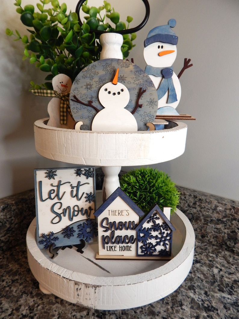 May include: A white tiered tray decorated with winter-themed wooden cutouts. The top tier features a snowman, a blue and white snowflake design, and a small green bush. The bottom tier features a sign that says "Let it Snow", two houses with the text "There's Snow Place Like Home", and a small green bush.